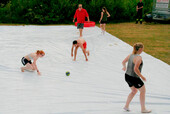 Hier sehen Sie, wie 2 Mannschaften auf einer großen weißen Plane das Spiel "Brennball" spielen. Dabei versuchen sie das Gleichgewicht zu halten, da die Oberfläche sehr rutschig ist.